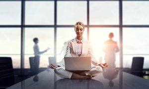 A woman sitting on a conference table with a laptop on her lap and in a meditation pose.